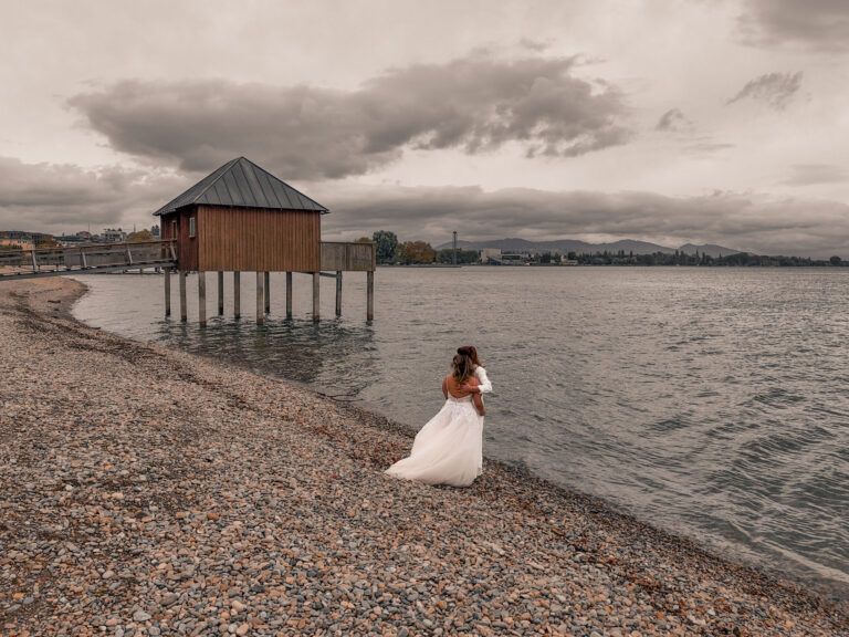 Hochzeitsfotograf Aalen – Romantische Hochzeitsaufnahme eines Brautpaares am Seeufer des Bodensees mit dramatischem Himmel. Stimmungsvoll inszenierte Hochzeitsfotografie mit natürlichen und emotionalen Momenten
